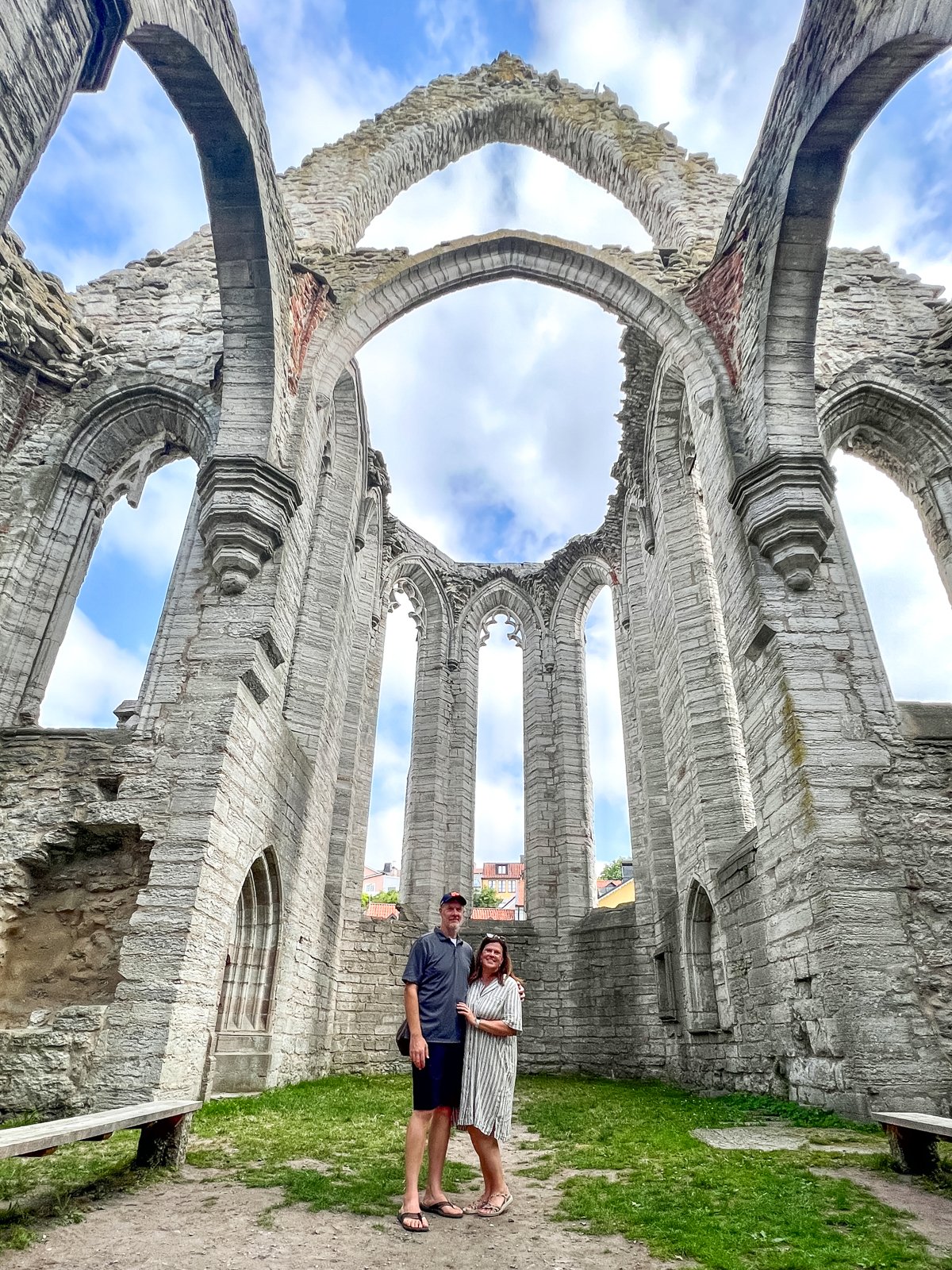 Two people stand under the arched ruins of a stone structure with a blue sky and clouds visible through the open ceiling.