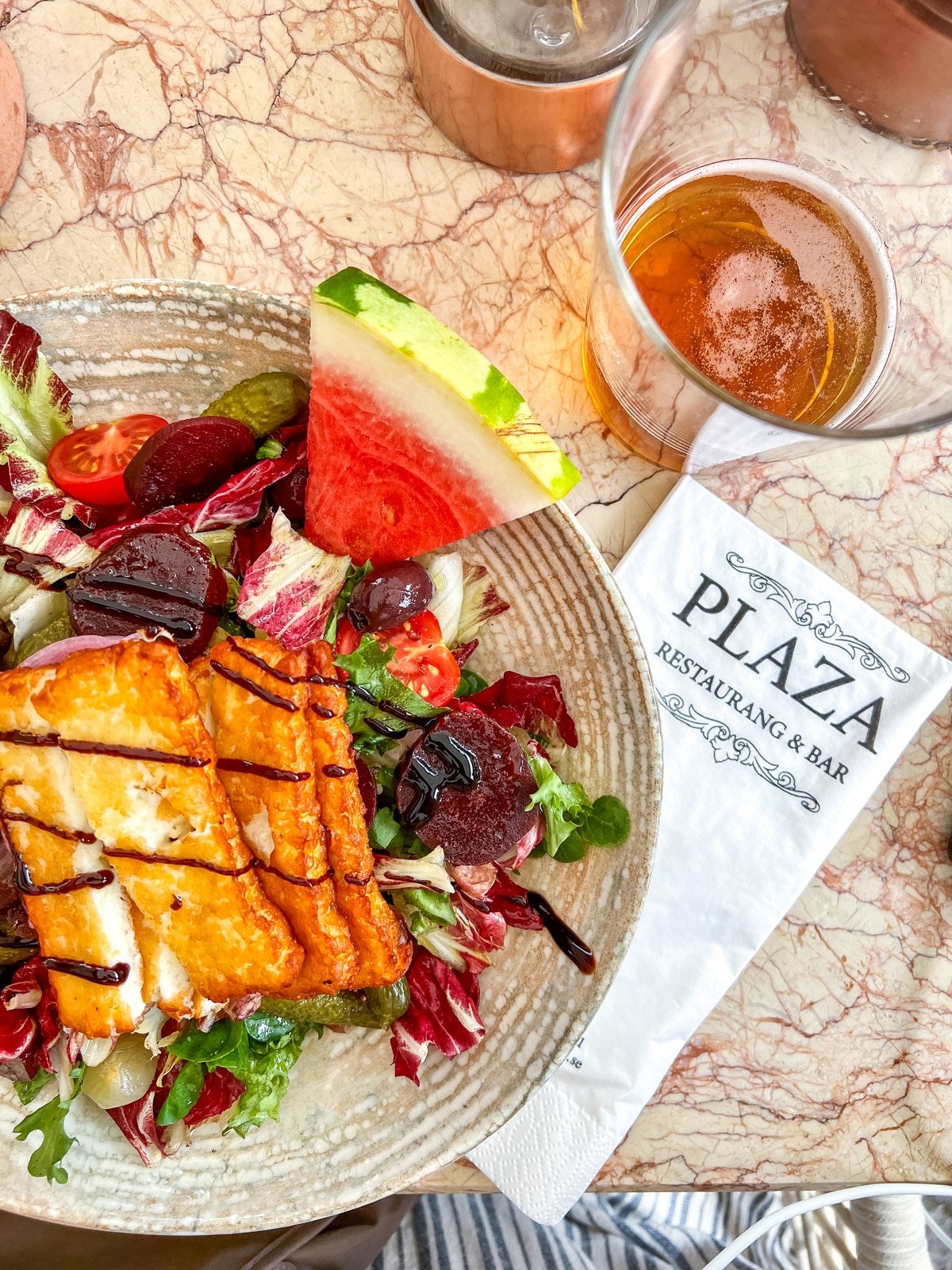 A plate with grilled cheese on a bed of mixed greens, beets, and watermelon, accompanied by a glass of beer on a marble table. A napkin with the text "PLAZA RESTAURANG & BAR" is beside the plate.