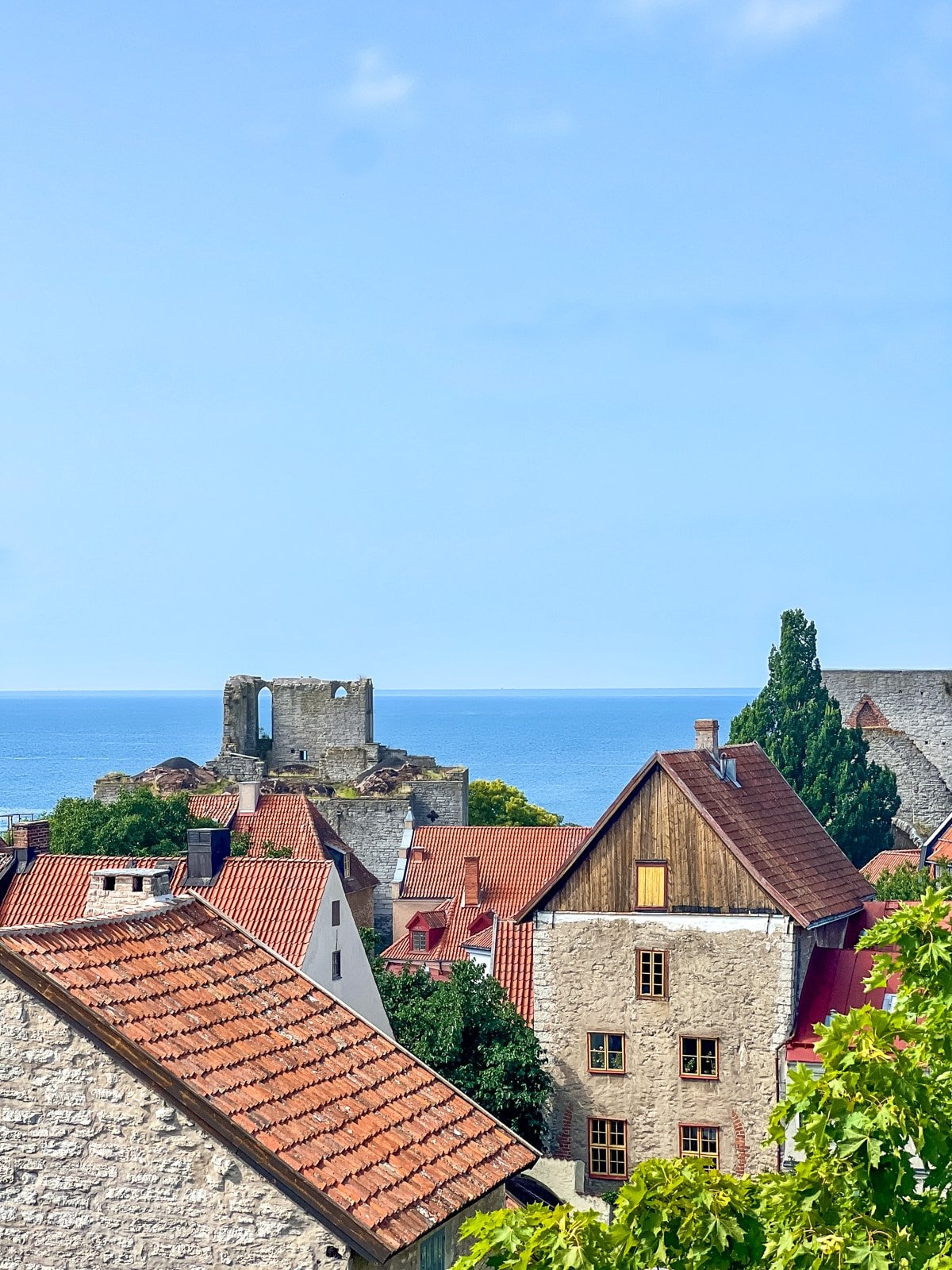 View of red-roofed houses with stone walls and a historic ruin by the sea under a clear, blue sky.