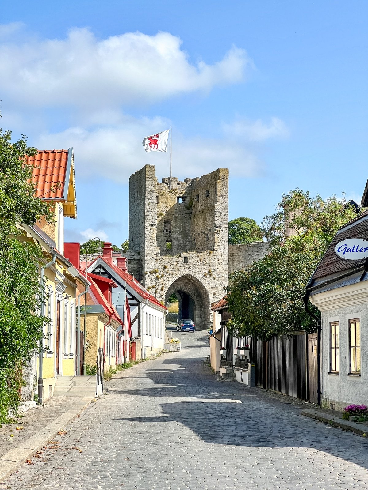 A historic stone gate stands at the end of a cobblestone street lined with quaint houses. A flag flies from the top of the gate under a blue sky.