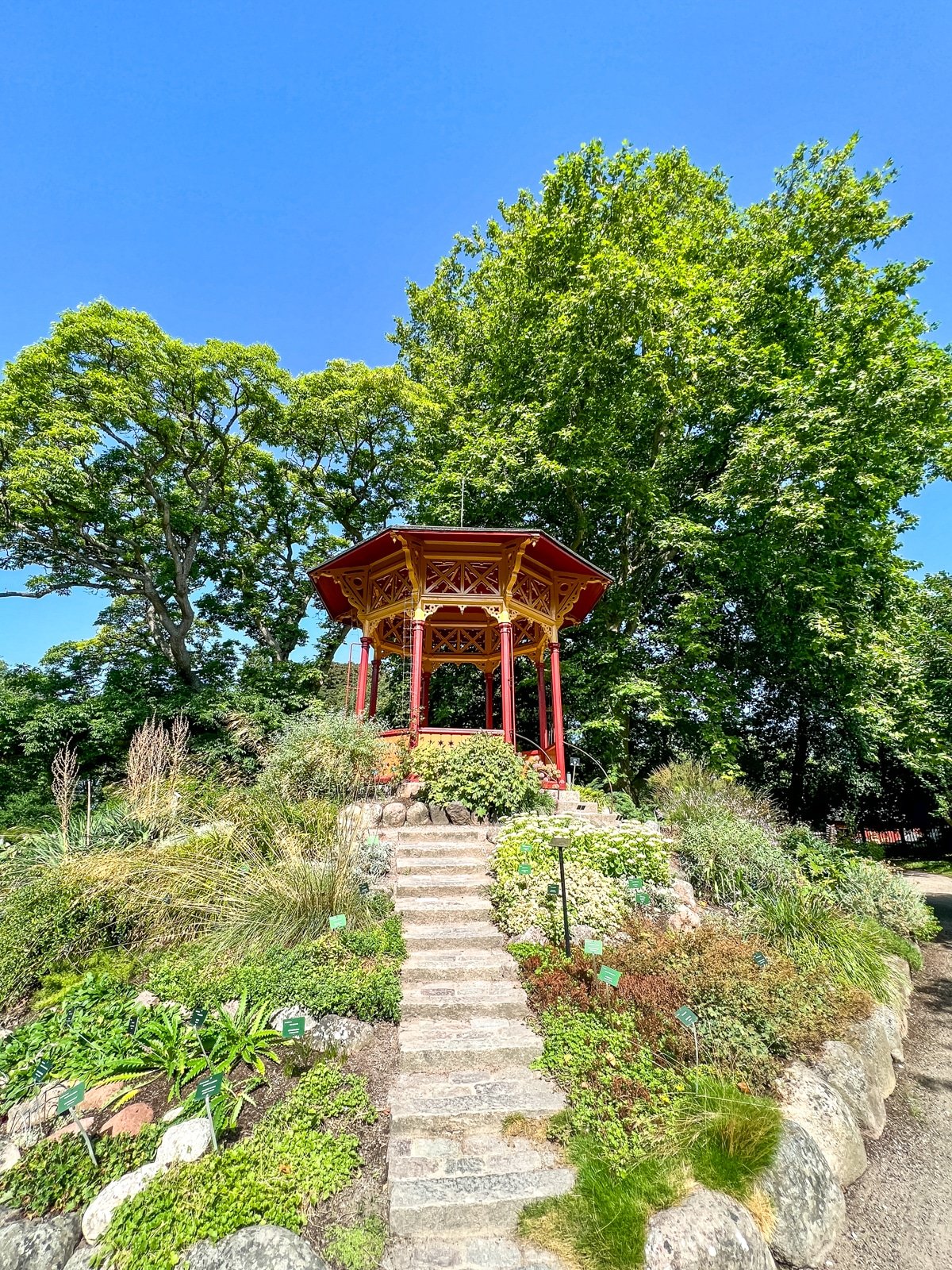 A red gazebo sits atop a small hill surrounded by lush greenery and plants, with stone steps leading up to it under a clear blue sky.