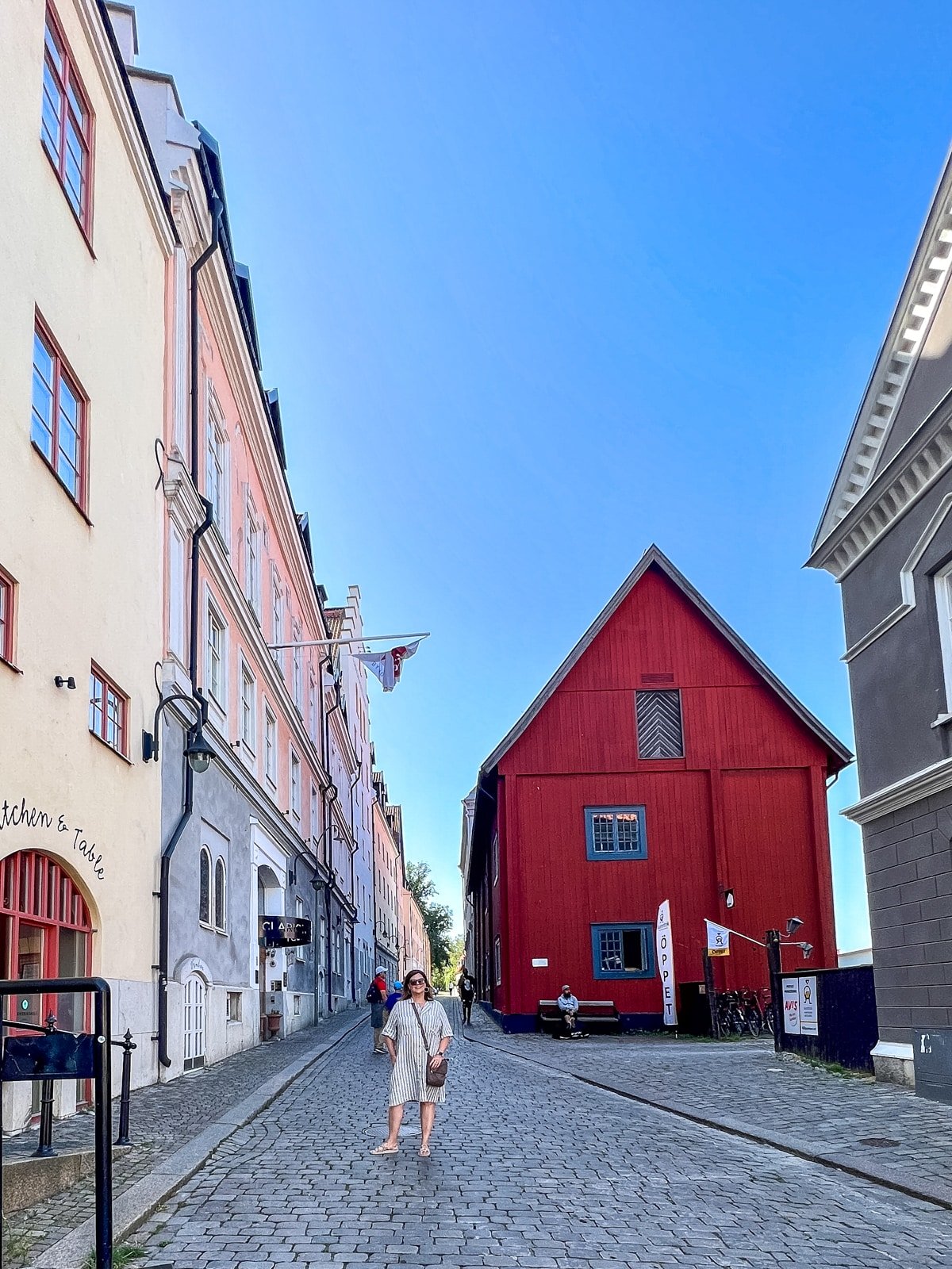 A person stands on a cobblestone street surrounded by colorful buildings, including a distinctive red wooden structure, under a clear blue sky.