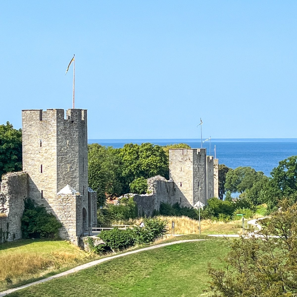 Stone towers and fortified walls of a historic structure stand amidst greenery with the ocean visible in the background under a clear blue sky.