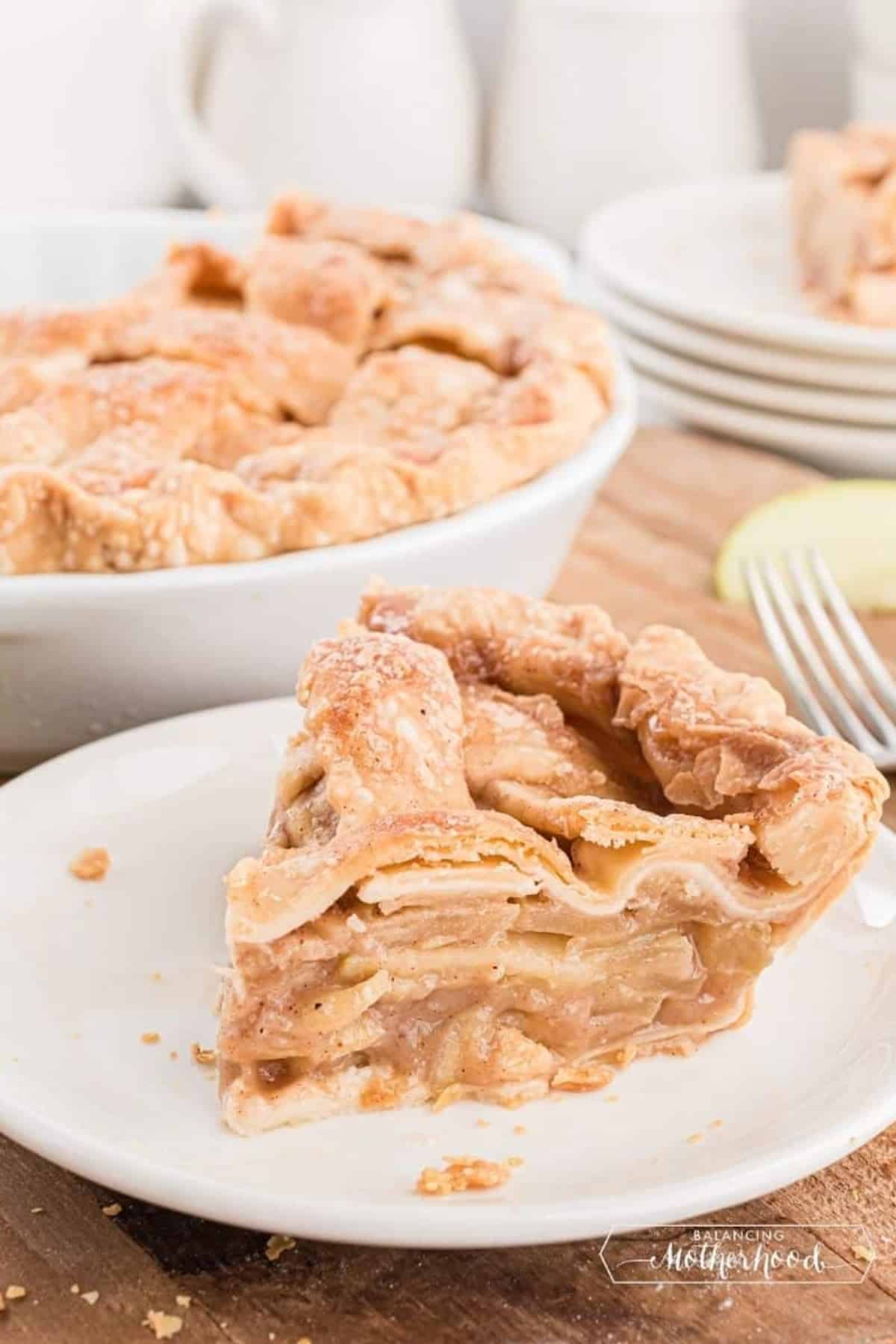 A slice of layered apple pie with a flaky golden crust is served on a white plate. The remaining pie is in a dish in the background.