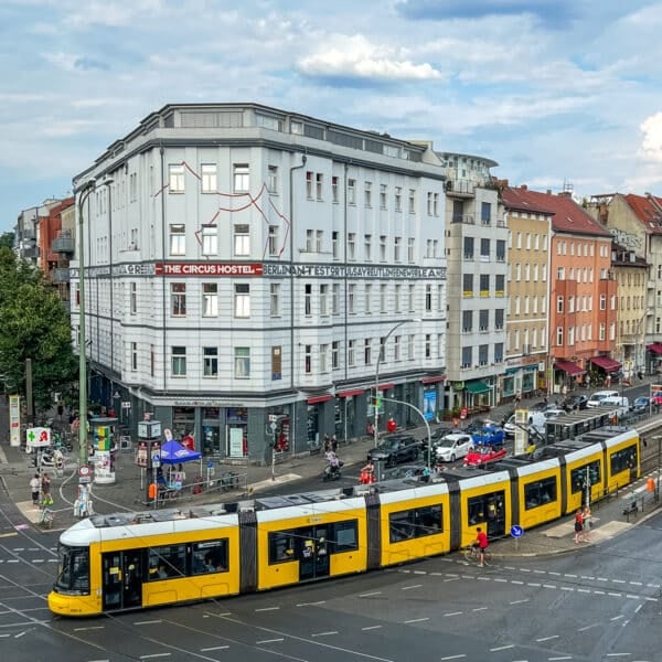 A yellow tram travels through a busy intersection in an urban area with multiple buildings, cars, and pedestrians.