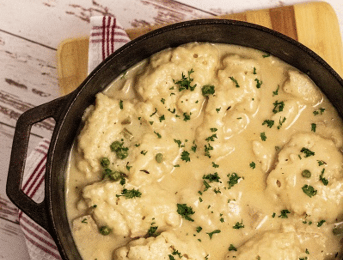 A cast iron skillet filled with creamy chicken and dumplings, garnished with parsley, on a wooden surface with a red and white striped cloth.