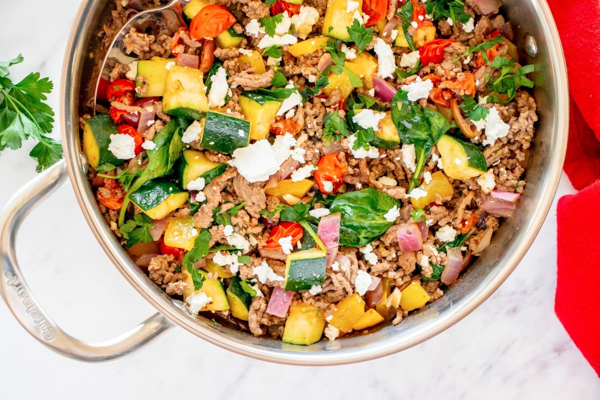 A skillet filled with cooked ground beef, zucchini, tomatoes, spinach, and crumbled feta cheese, garnished with parsley.