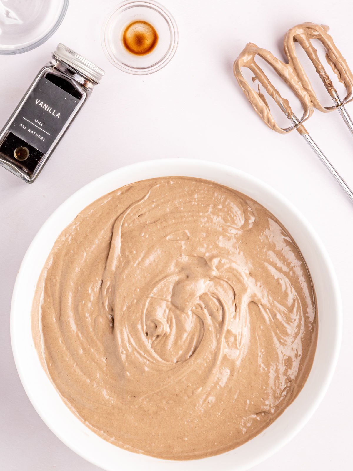 A mixing bowl with chocolate cake batter, a bottle of vanilla extract, two beater attachments with batter, and a small dish with liquid are arranged on a white surface.