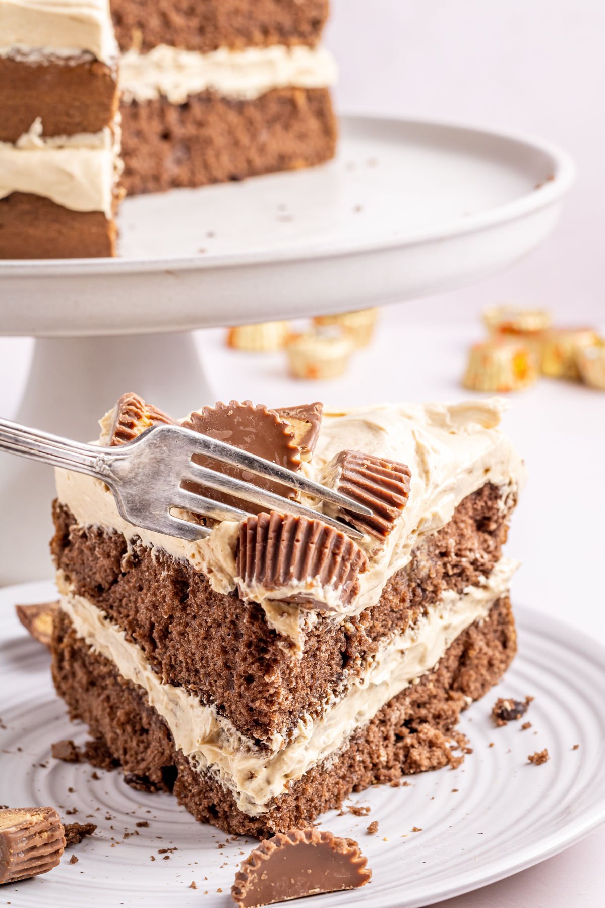 A slice of chocolate cake with layers of frosting and topped with peanut butter cups sits on a plate. A fork rests on the slice, and more cake is on the stand in the background.