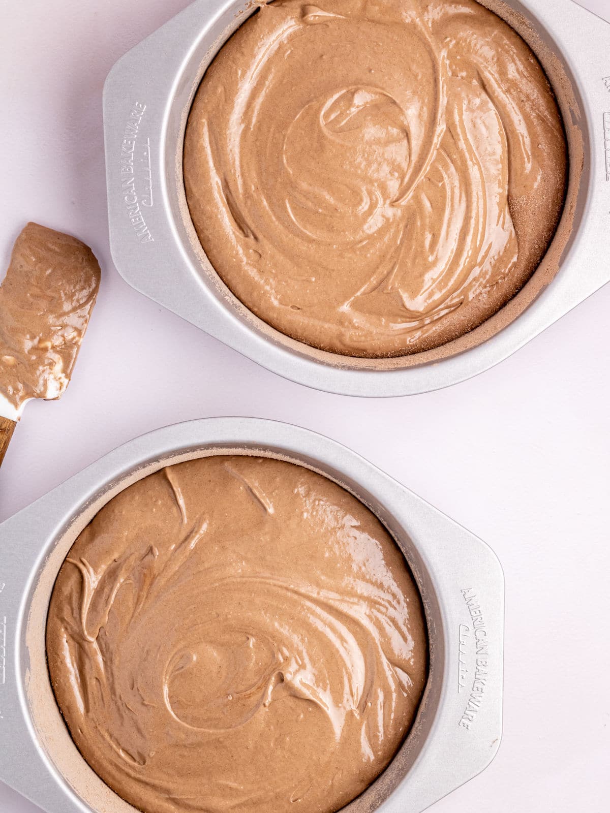 Two round baking pans filled with swirled chocolate cake batter sit on a light surface, beside a spatula with remaining batter.