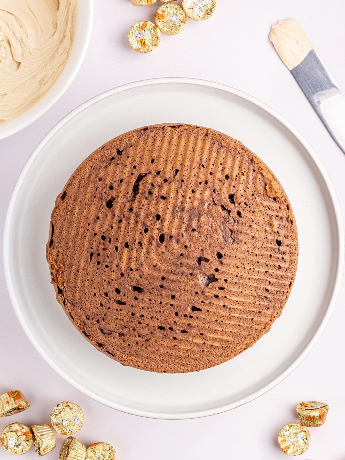 A chocolate cake on a round white plate, surrounded by unwrapped gold foil chocolates. A bowl of frosting and a knife with frosting are nearby.