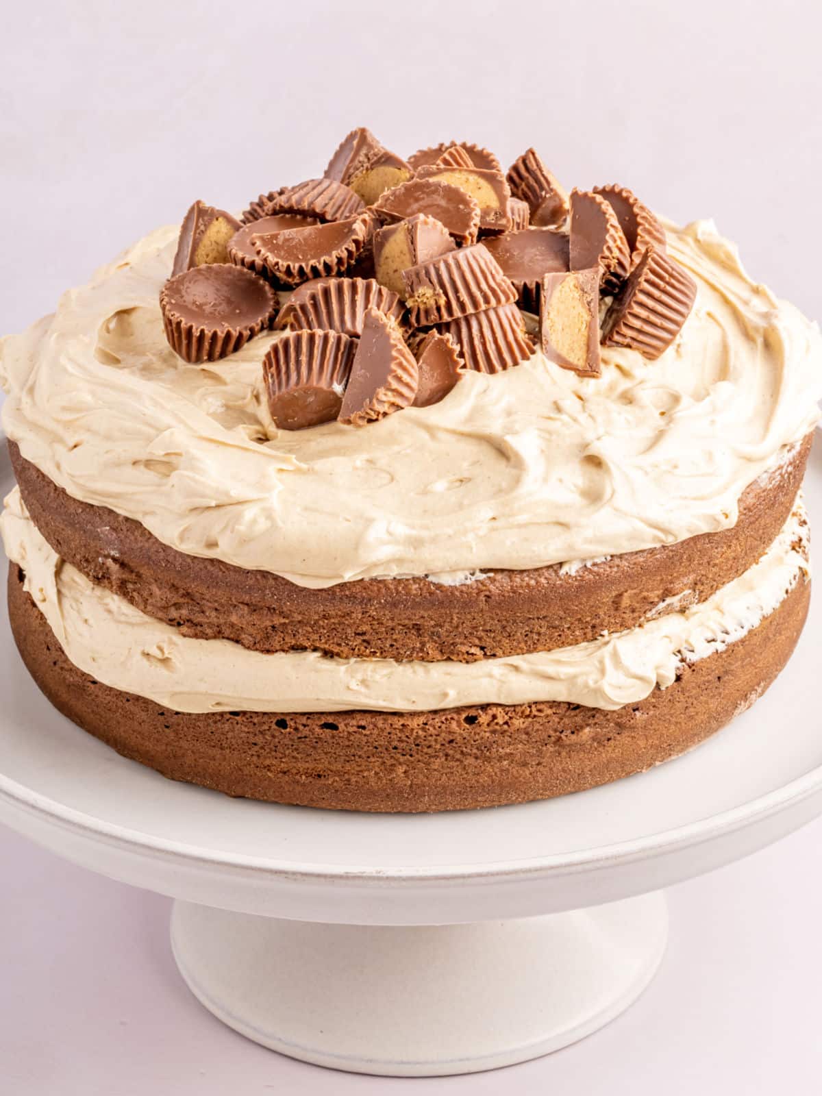 Two-layer chocolate cake with light brown frosting, topped with halved peanut butter cups, on a white cake stand.