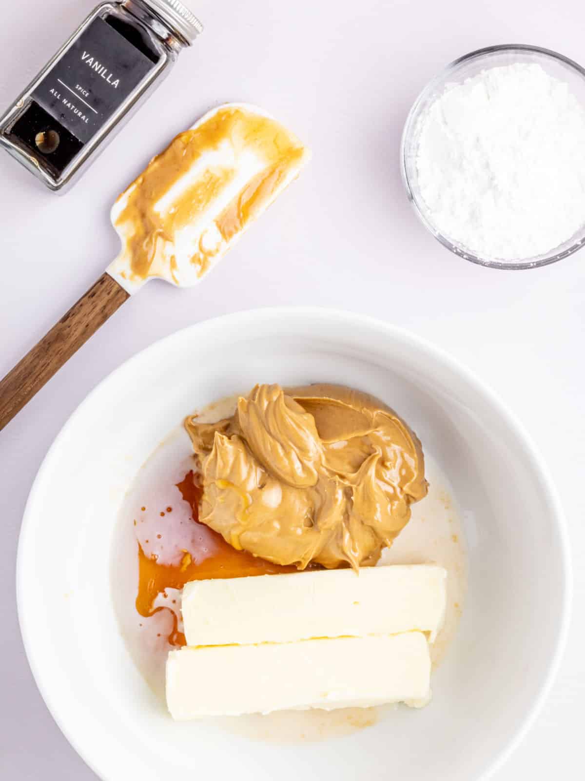 A bowl with peanut butter, butter sticks, and vanilla, next to a spatula with remnants, a vanilla extract bottle, and a glass bowl of powdered sugar.