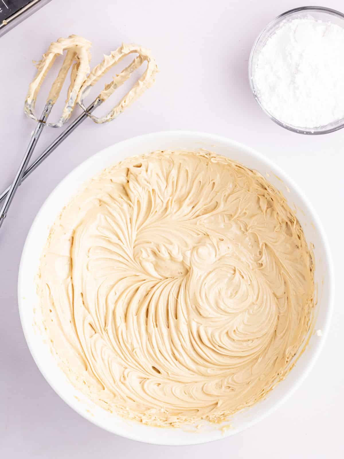 A mixing bowl filled with creamy light brown frosting is on a white surface next to beaters and a small bowl of powdered sugar.