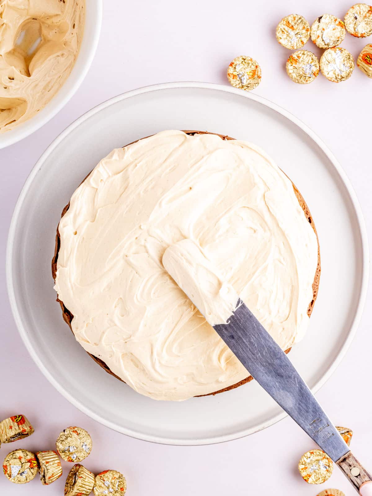 A cake being frosted with buttercream on a plate, surrounded by unwrapped peanut butter cups.