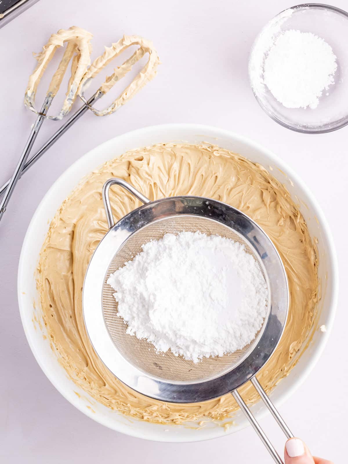 A bowl of brown batter with powdered sugar being sifted over it. Two beaters rest on the edge, and a bowl of powdered sugar is placed nearby.