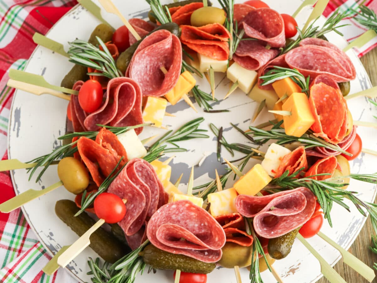 A charcuterie wreath with salami, pepperoni, cheese cubes, cherry tomatoes, olives, gherkins, and rosemary sprigs on skewers arranged in a circular pattern.