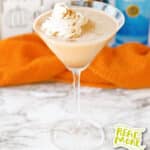 Pumpkin pie martini in a martini glass topped with whipped cream and cinnamon, set on a marble surface with bottles and an orange cloth in the background.