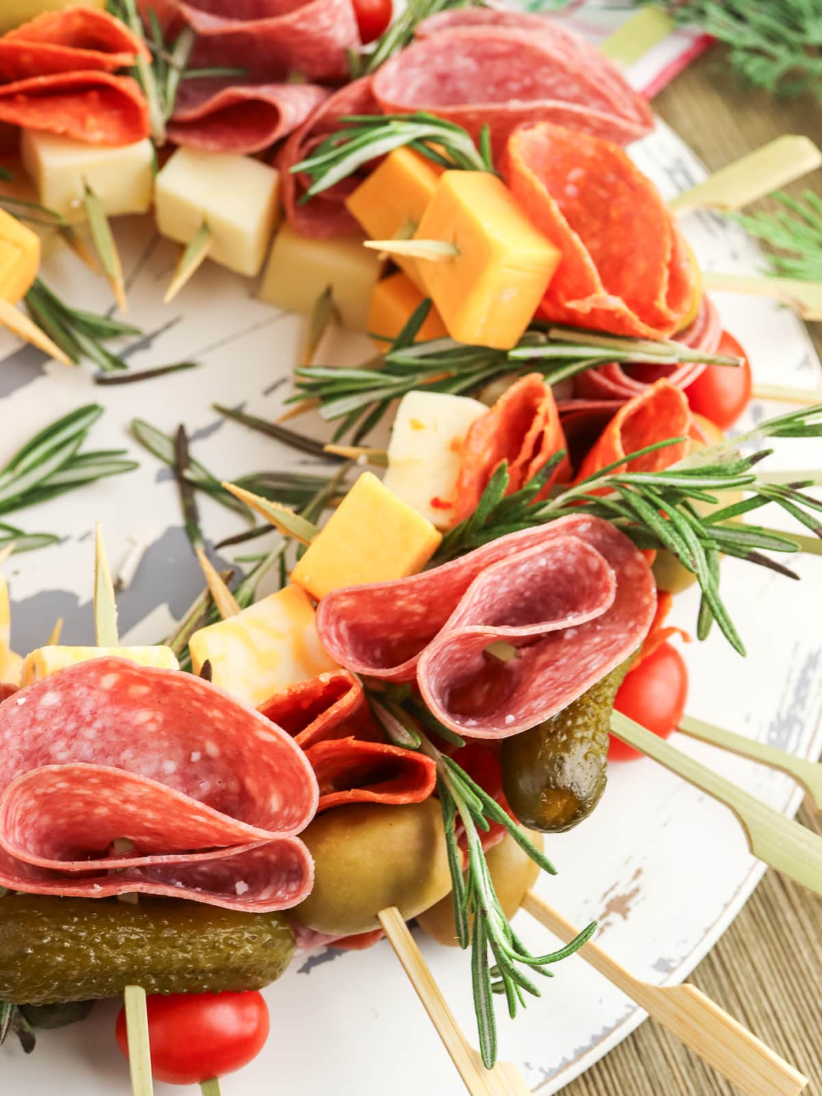 A festive meat and cheese wreath with salami, cheddar, gherkins, cherry tomatoes, and rosemary skewers arranged on a white platter.