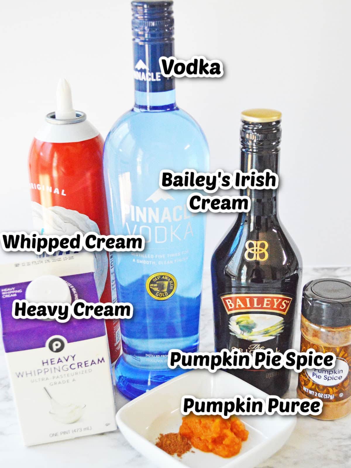 Ingredients for a cocktail: vodka, Bailey's Irish Cream, whipped cream, heavy cream, pumpkin pie spice, and pumpkin puree displayed on a countertop.