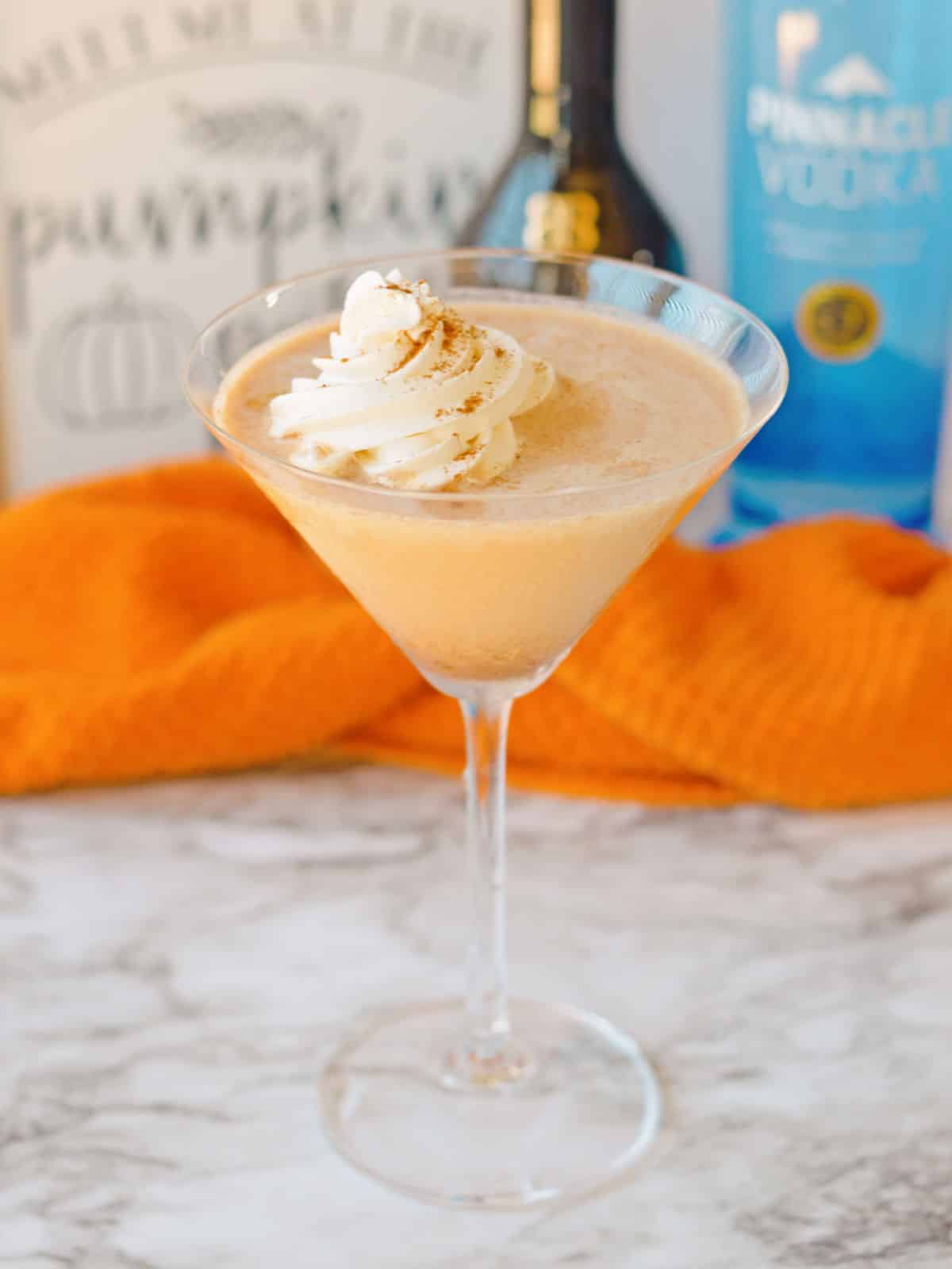 A pumpkin spice martini with whipped cream in a glass, set on a marble surface with an orange cloth and two liquor bottles in the background.