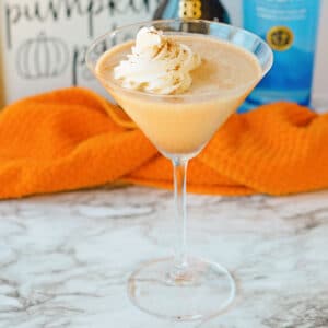 A pumpkin spice martini in a glass with whipped cream topping, set on a marble surface. An orange cloth and bottles are blurred in the background.