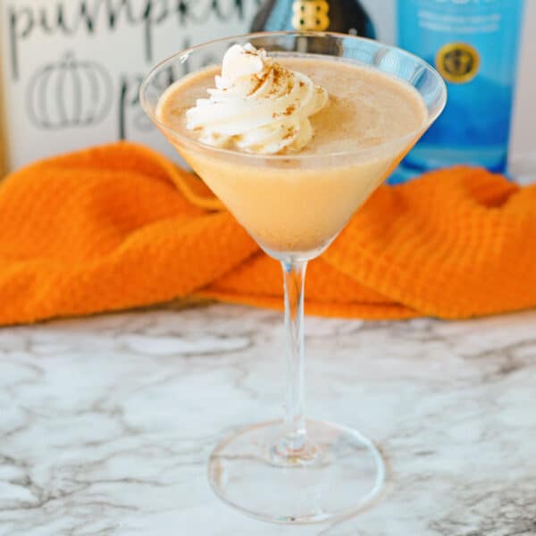 A pumpkin spice martini in a glass with whipped cream topping, set on a marble surface. An orange cloth and bottles are blurred in the background.