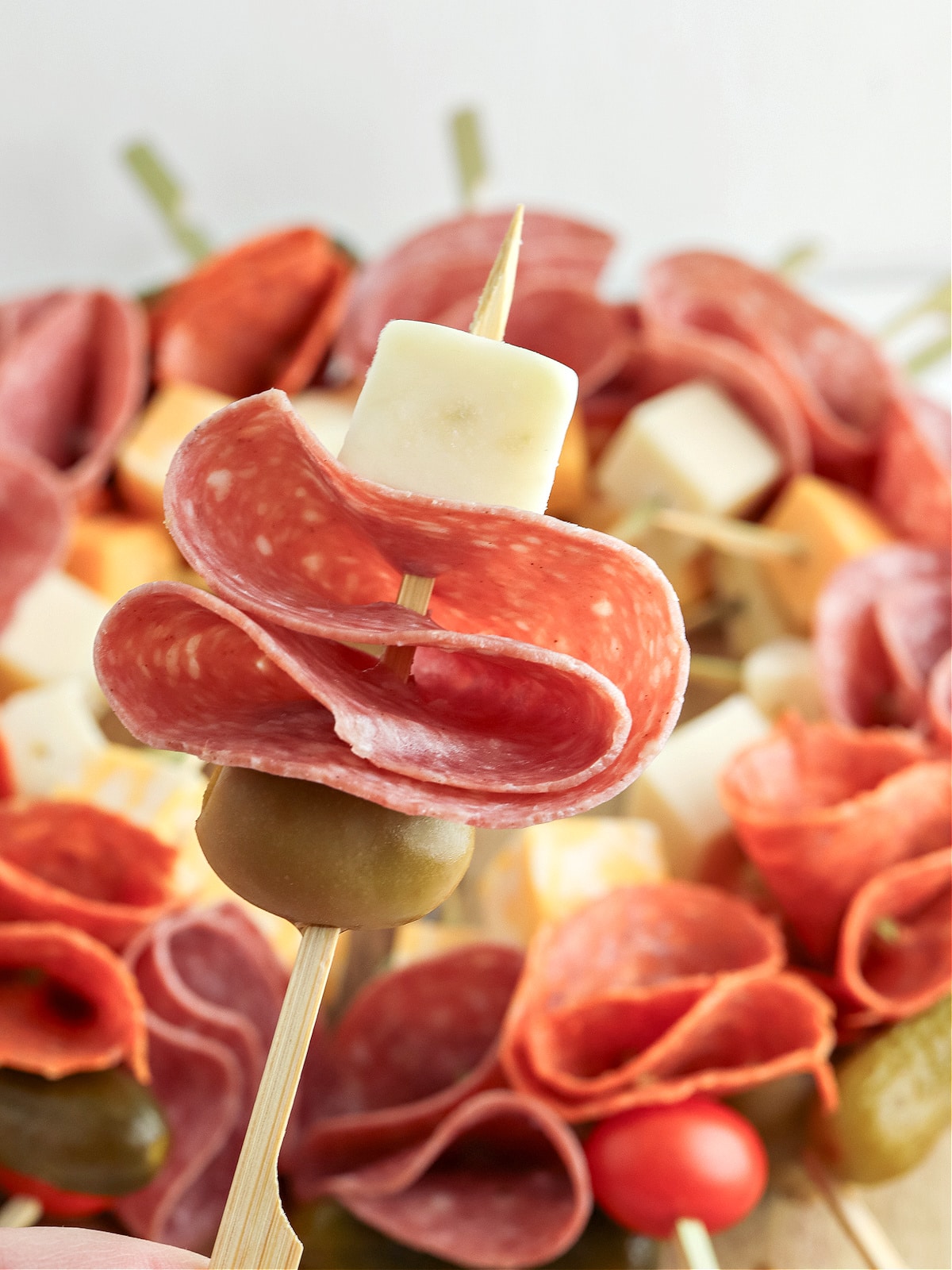 A skewered appetizer featuring salami, cheese, and an olive is held in focus, with assorted deli meats and cheeses in the background.