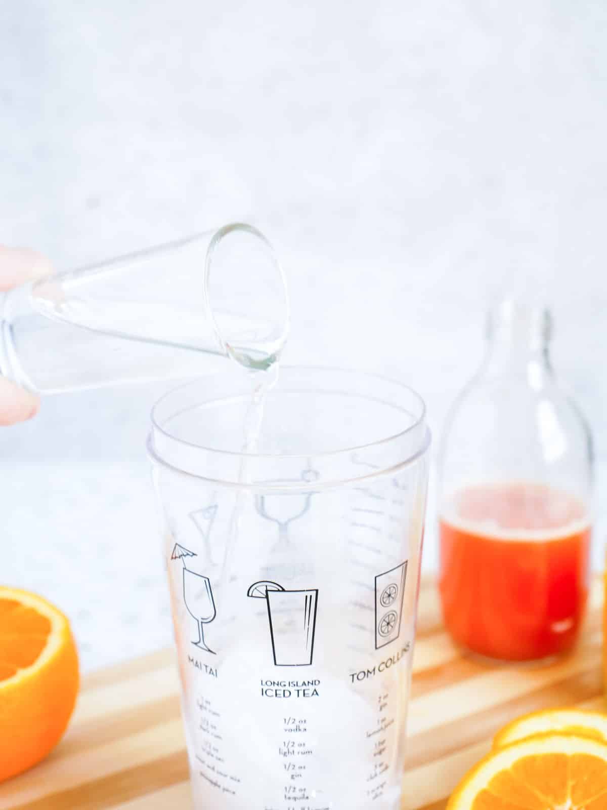 A shot glass pouring liquid into a cocktail shaker, with sliced oranges and a bottle of red juice on a wooden surface.