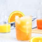 A glass of orange juice with ice and a slice of orange on the rim, placed on a marble surface. A carafe and another sliced orange are in the background.