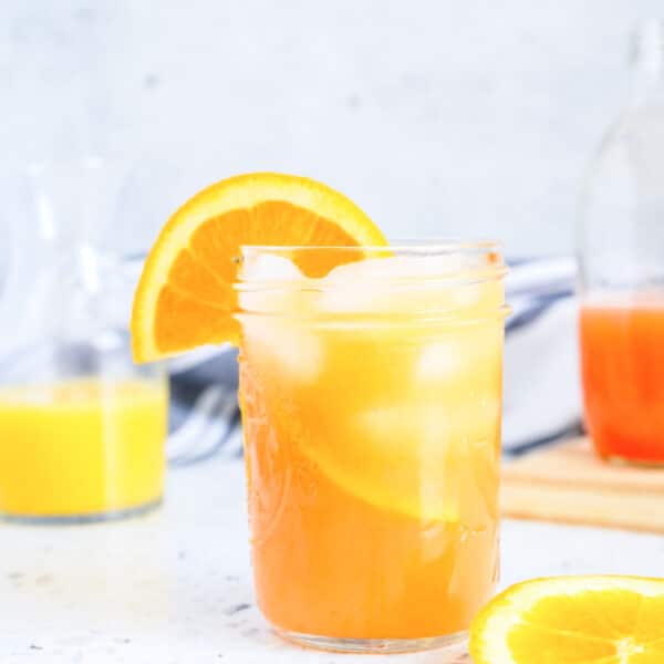 A glass of orange juice with ice and a slice of orange on the rim, placed on a marble surface. A carafe and another sliced orange are in the background.