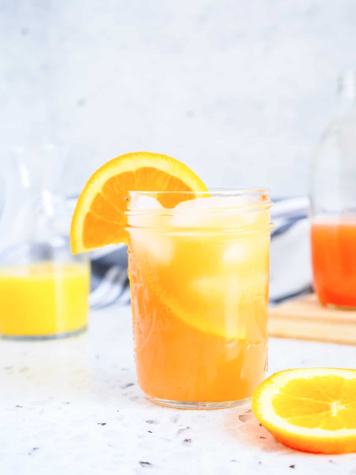 A glass of orange juice with ice and a slice of orange on the rim, placed on a marble surface. A carafe and another sliced orange are in the background.