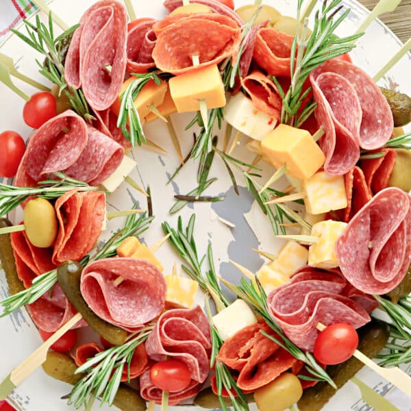 A platter with a wreath arrangement of salami slices, cheese cubes, cherry tomatoes, olives, pickles, and rosemary sprigs on skewers.