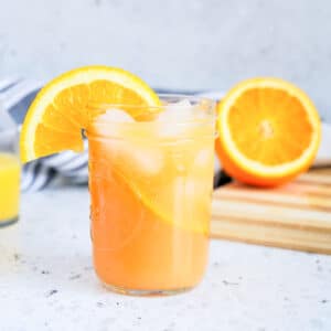 A mason jar filled with iced orange drink, garnished with an orange slice, sits on a counter next to a half-sliced orange and a striped cloth.