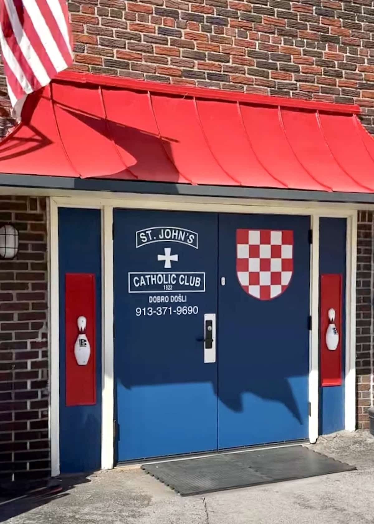 Entrance of St. John's Catholic Club featuring blue doors with a white cross and checkered shield. Red awning above and brick walls surrounding, with a visible phone number.