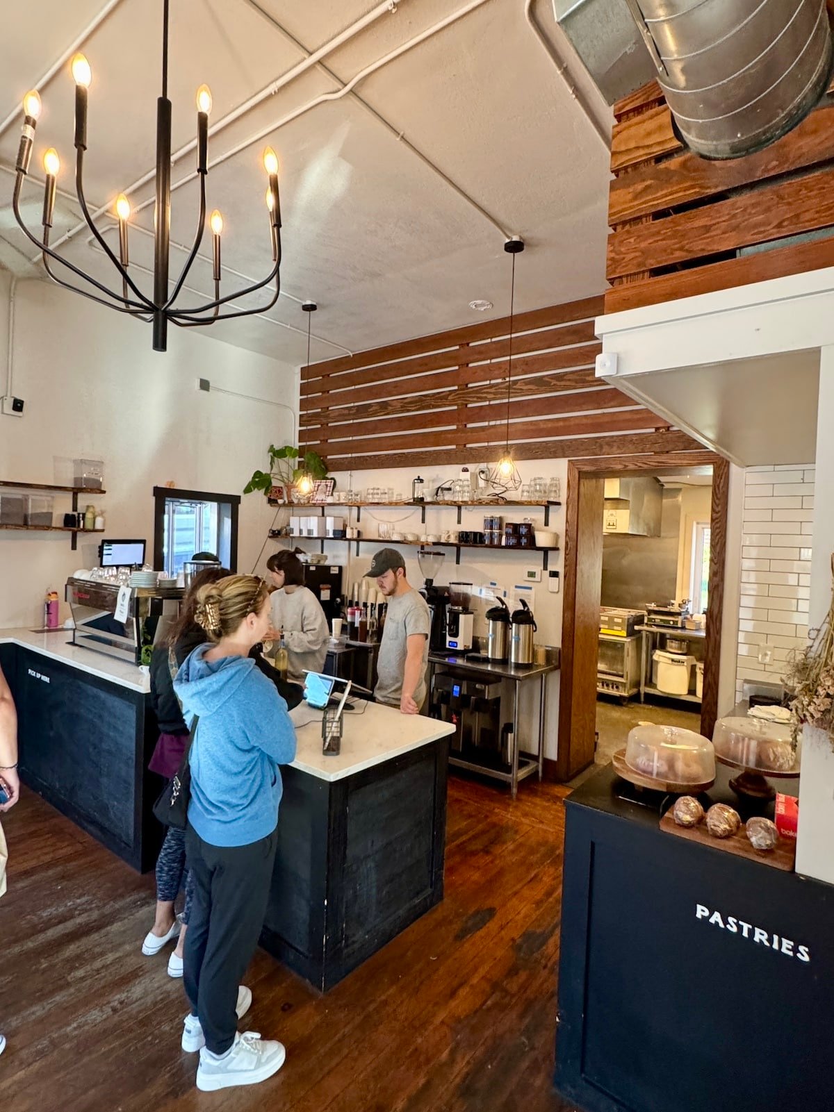 Customers order at a cafe counter with a menu board above. Bread and pastries are displayed nearby. An employee assists patrons under hanging lights.