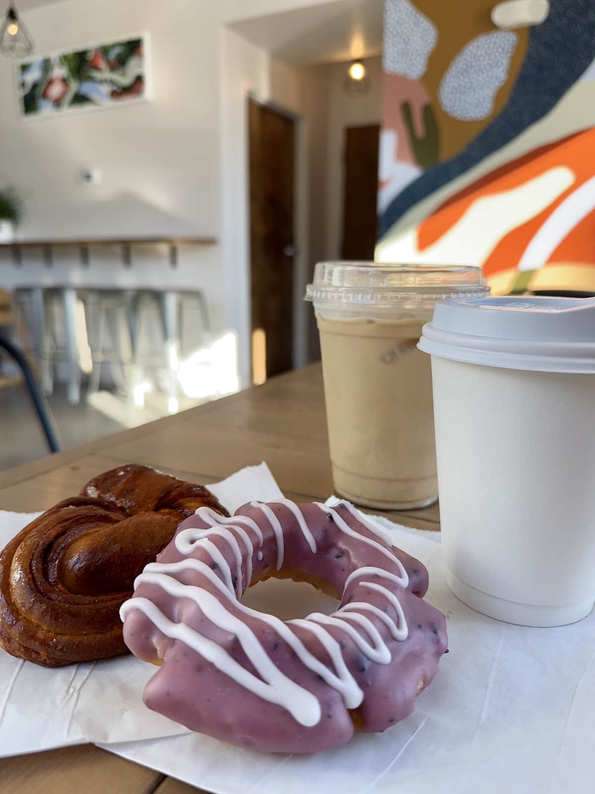 Two pastries on a table, one with pink icing and white drizzle. Next to them are two drinks in cups, one with a clear lid. Background features a colorful mural and seating area.