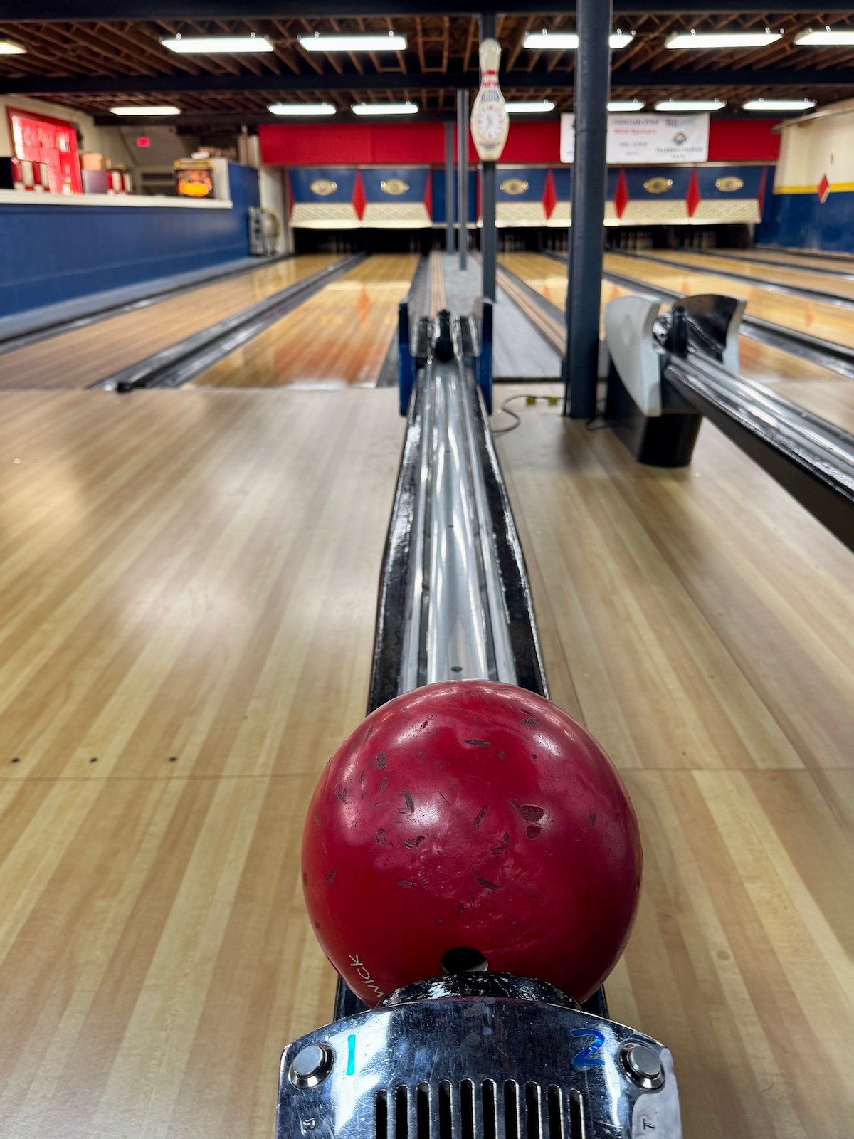 Red bowling ball on a ball return at a bowling alley, with lanes and pins in view.