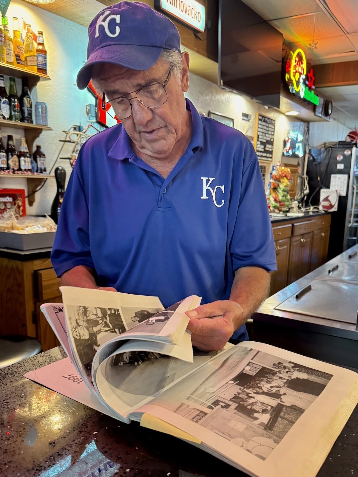 Man in a "KC" shirt reads a book at a bar counter, surrounded by bottles and lit signs.