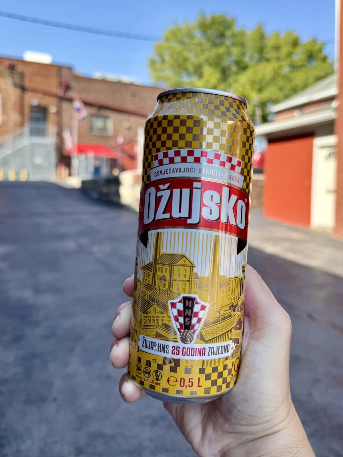 A hand holding a can of Ožujsko lager against a blurred urban background. The can features special commemorative packaging.