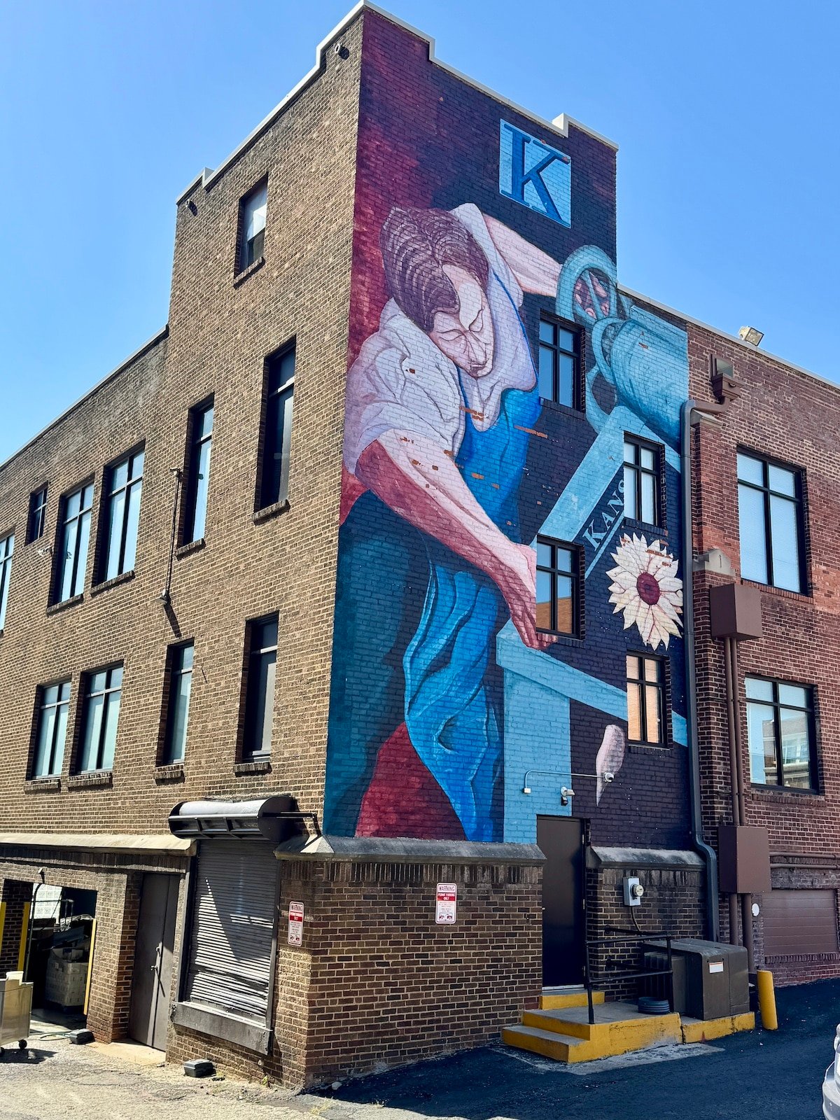 Mural on a brick building depicting a person working with machinery under a large letter "K".