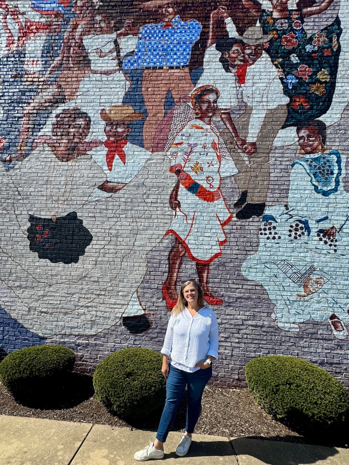 A woman stands in front of a colorful mural depicting people in traditional attire dancing.