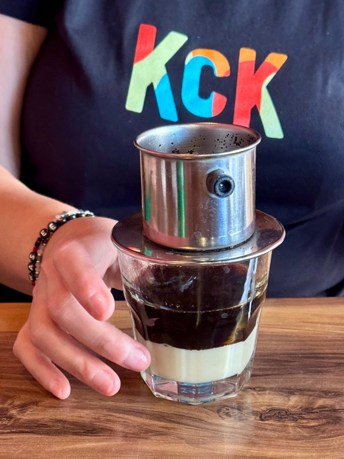 A Vietnamese drip coffee maker is placed on a glass containing sweetened condensed milk. A person in a black shirt with colorful letters holds the glass.