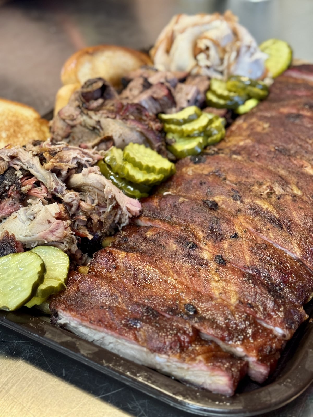 A platter with sliced pork ribs, pulled pork, and chicken. Accompanied by sliced pickles and bread rolls.