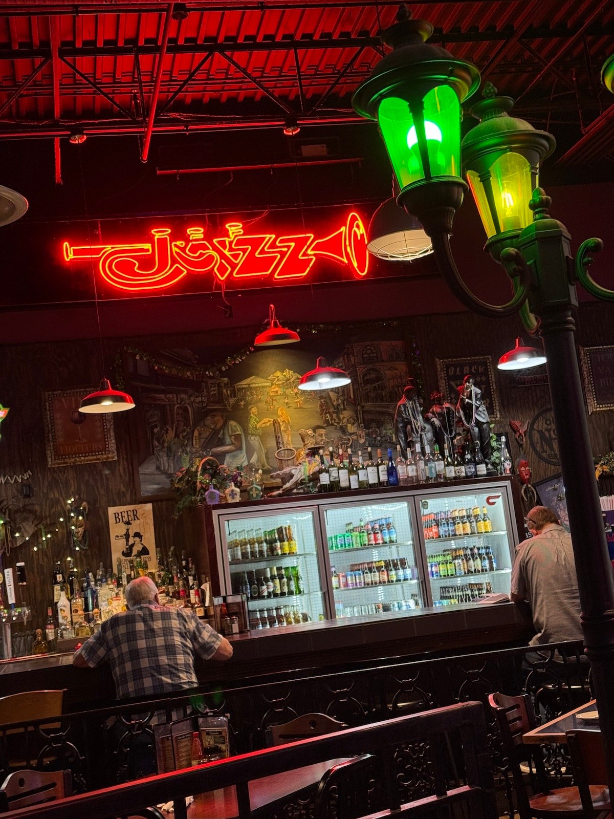 A cozy bar with two people seated at the counter. A neon "Jazz" sign glows above, and a lit green lamp is in the foreground, creating a relaxed ambiance.