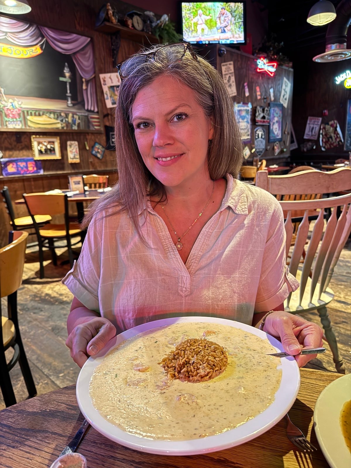 A woman with long hair smiles while holding a large plate of creamy soup with a mound of rice in the center, seated in a restaurant.