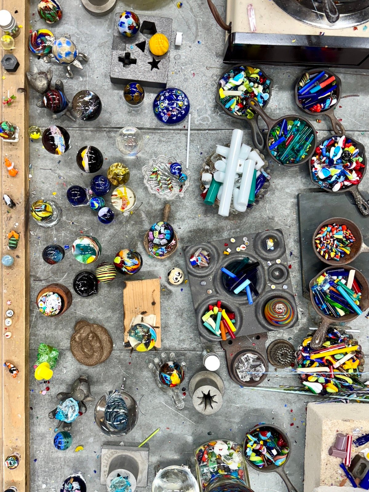 An overhead view of a cluttered workspace with colorful glass pieces, tools, and rods arranged on a table, indicating glassblowing activities.