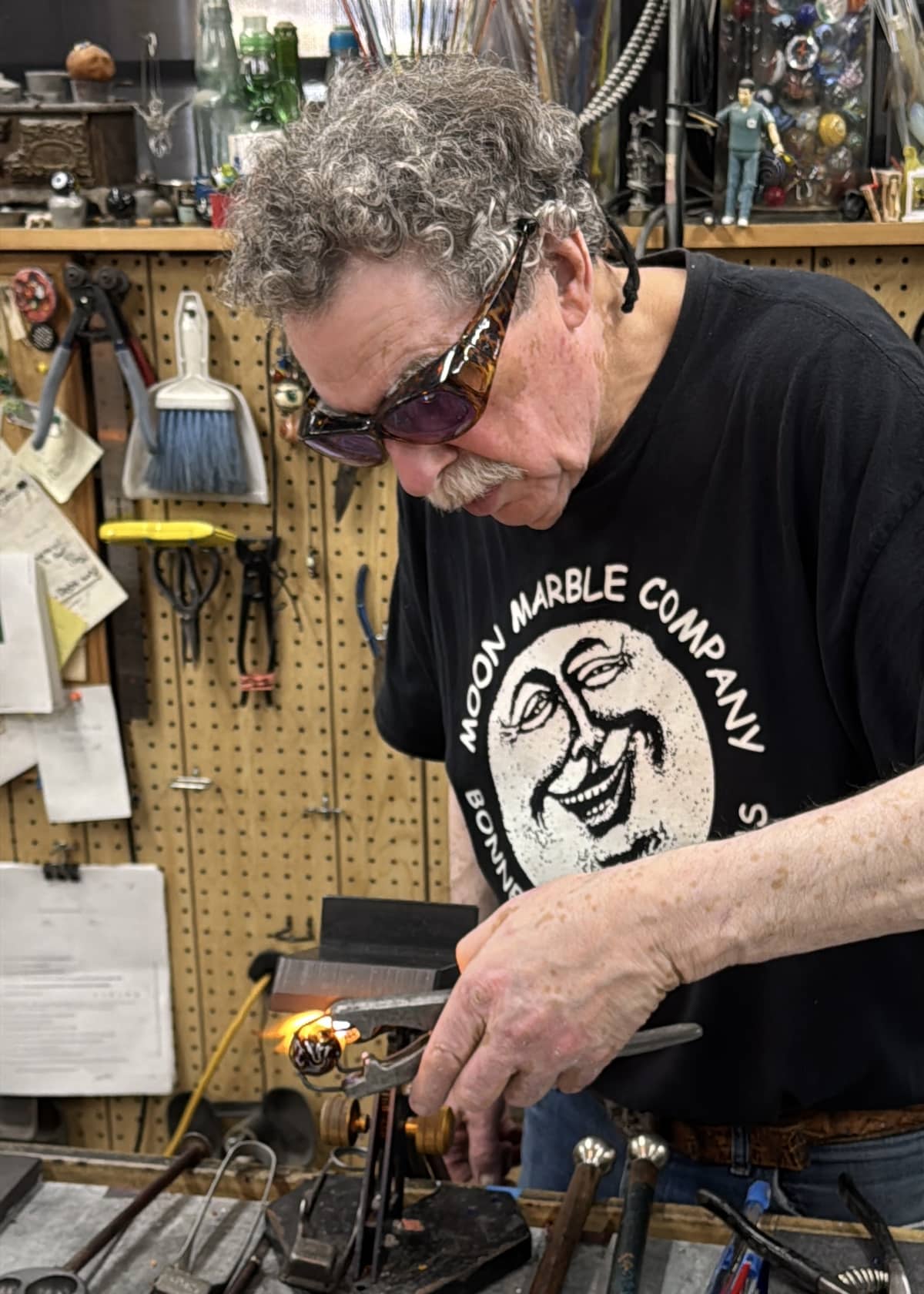 An artisan with curly hair and sunglasses works on a glass creation using pliers in a workshop filled with tools and hanging items.