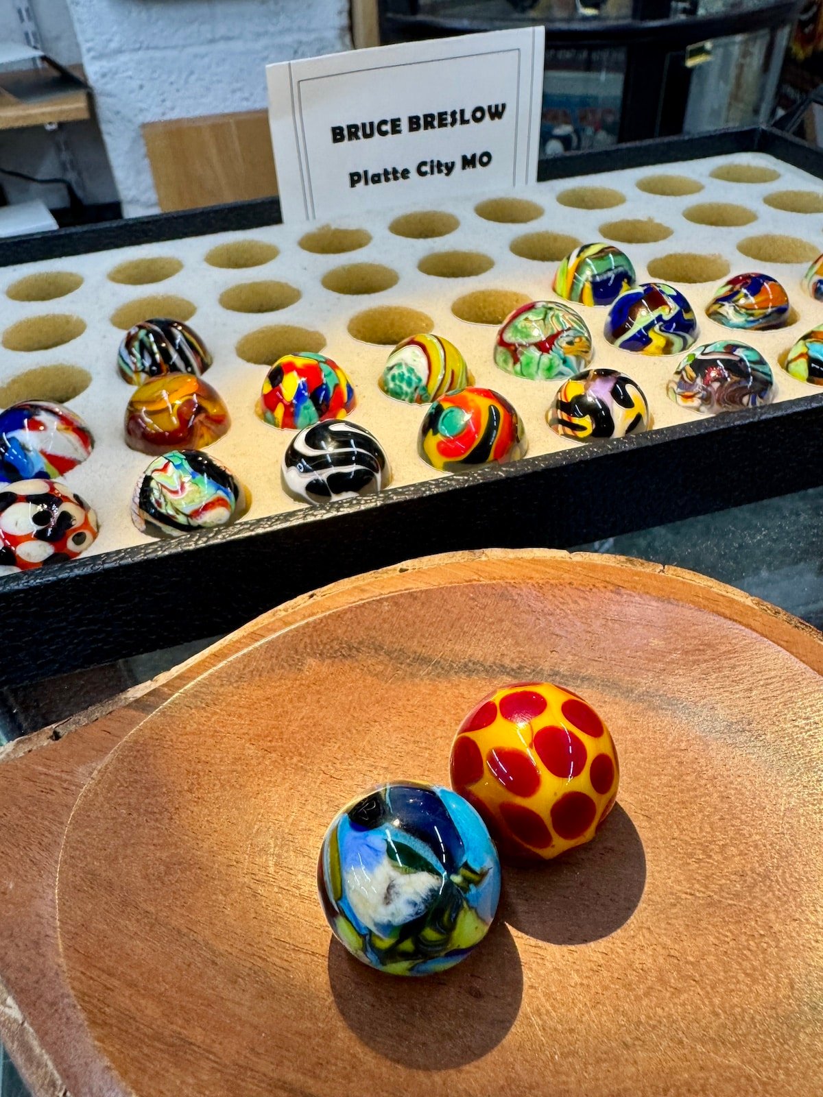 Display of colorful, patterned marbles on a wooden tray and a holder. Sign reads "Bruce Breslow, Platte City MO.