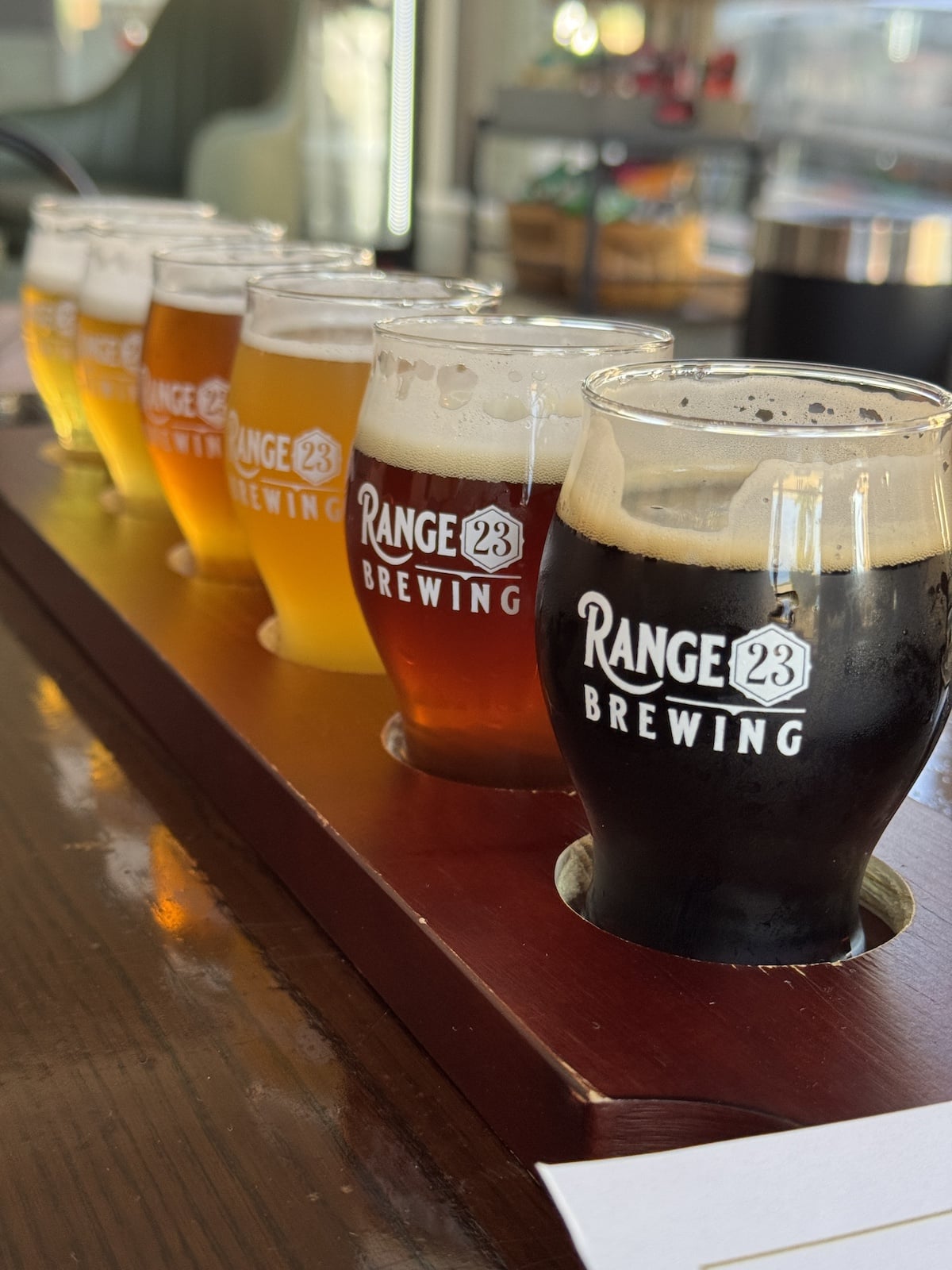 A flight of five assorted craft beers in small glasses on a wooden tray labeled "Range 23 Brewing.